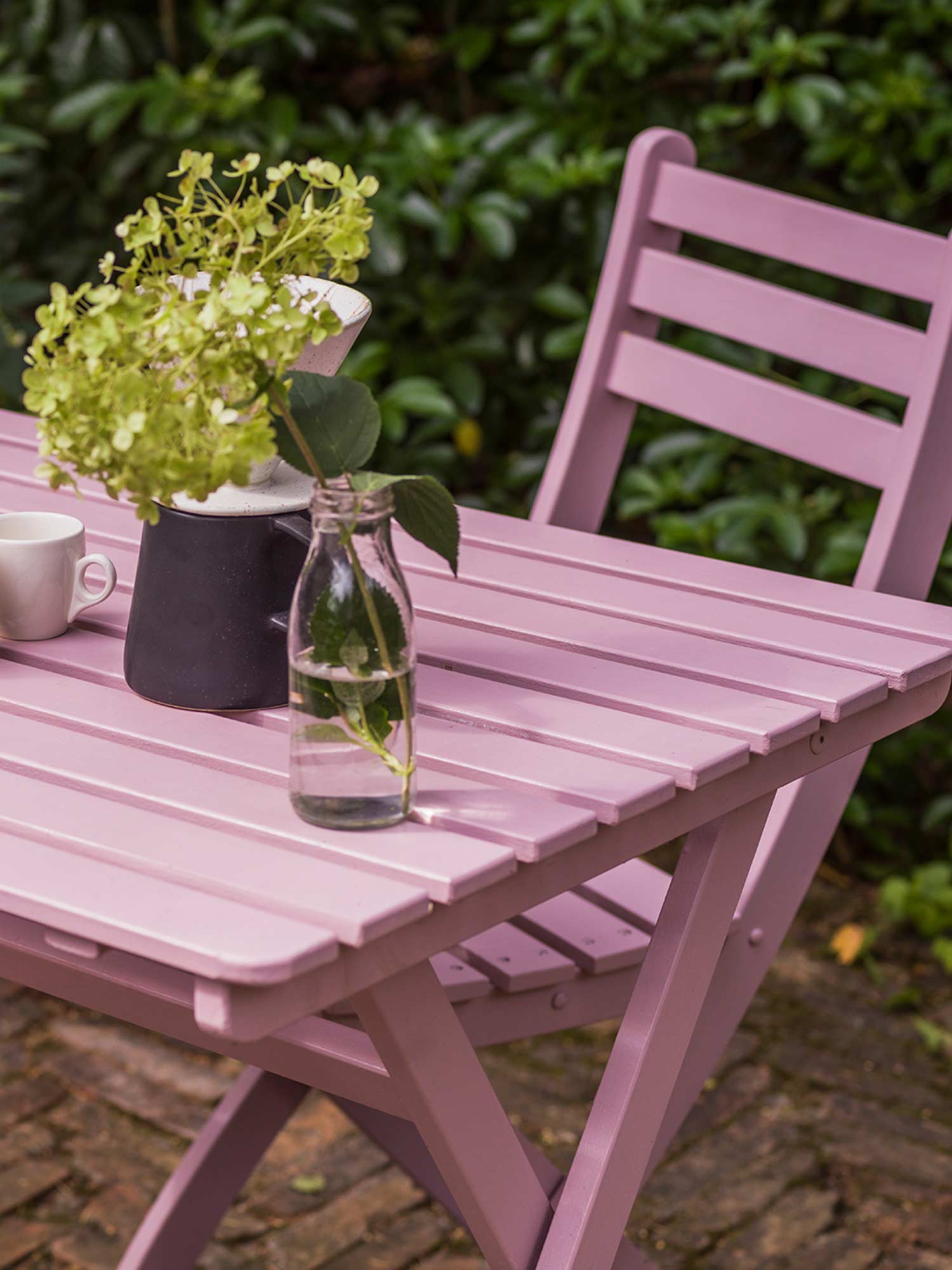 Gartenmöbel in Cinder Rose No. 246 von Farrow & Ball. Wildromantisches Pink auf Holztisch und Stuhl vor grünem Strauch.